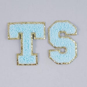 May include: Two embroidered patches, a "T" and an "S", in light blue with gold glitter trim. The letters have a textured, fuzzy appearance. The patches are likely for clothing or accessories.