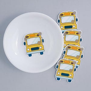 May include: Yellow school bus stickers with floral accents. The stickers are arranged on a white plate against a gray background. The bus design includes blue wheels and a blue stripe across the front.