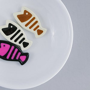 May include: Three ceramic fish figurines on a white plate. The fish are brown, black and white, and pink and black.