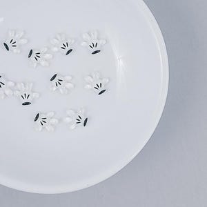 May include: A white ceramic bowl filled with small, white, Mickey Mouse-style hand-shaped decorations with black accents. The bowl is set against a light gray background.