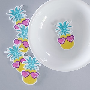 May include: Five yellow pineapple stickers with blue leaves and pink heart-shaped sunglasses. The stickers are on a white surface.