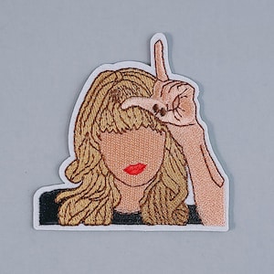 Puede incluir: Parche bordado con una ilustración estilizada de una persona con cabello rubio, labios rojos y una mano haciendo un gesto de perdedor. El parche tiene un borde blanco y una parte superior negra.