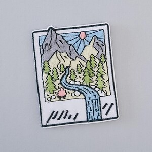 May include: An embroidered iron-on patch featuring a colourful illustration of a mountain range, a river, and a sun. The image is framed by a white border with a black outline and the word "//" at the bottom.