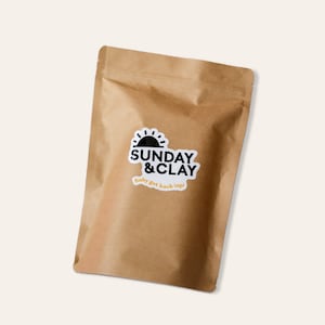 May include: A brown paper bag with a white sticker that says "SUNDAY & CLAY" with a black sun graphic and the text "Baby got back (up)".