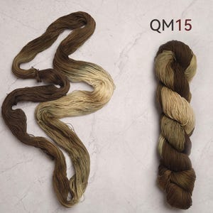 Peut inclure: Deux &eacute;cheveaux de fil de couleur marron et beige. L'un est dispos&eacute; en vrac, montrant des variations de couleur. L'autre est enroul&eacute; serr&eacute;, affichant un d&eacute;grad&eacute; du marron fonc&eacute; au beige. Le texte "QM15" est en haut &agrave; droite.