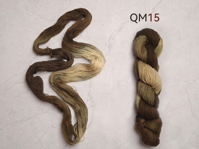 Peut inclure: Deux &eacute;cheveaux de fil de couleur marron et beige. L'un est dispos&eacute; en vrac, montrant des variations de couleur. L'autre est enroul&eacute; serr&eacute;, affichant un d&eacute;grad&eacute; du marron fonc&eacute; au beige. Le texte "QM15" est en haut &agrave; droite.