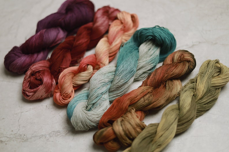 Six braided skeins of embroidery thread arranged in a diagonal row. The colours include deep purple, dark red, light peach, teal and white gradient, burnt orange, and olive green. Each skein features a soft, lustrous, and fine texture.
