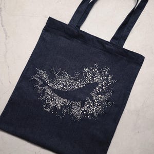 Pode incluir: Uma sacola tote de ganga azul escura com um design de baleia branca. A baleia é cercada por pontos brancos. A sacola tem duas alças de ganga a condizer.
