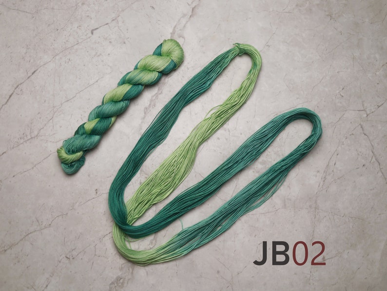 Op de afbeelding: Twee bollen groene borduurgaren, &eacute;&eacute;n bol is gedraaid en de andere ligt plat. De bollen liggen op een grijs marmeren oppervlak. De tekst "JB02" is op het oppervlak gedrukt.