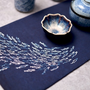 May include: A dark blue placemat with a design of white fish swimming in a school. The placemat is made of a soft, textured fabric and has a subtle sheen.