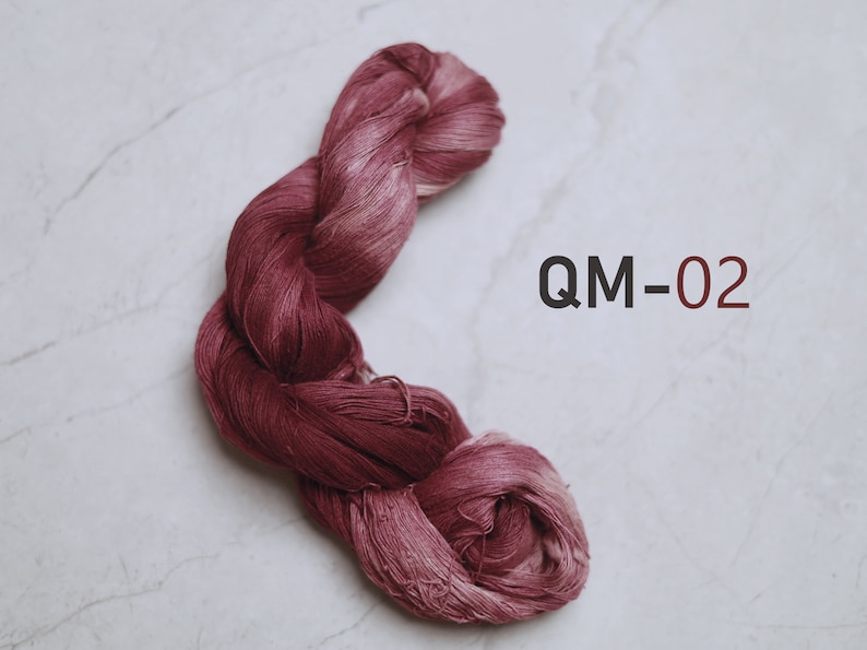 Puede incluir: Una madeja trenzada de hilo color burdeos profundo con un brillo sutil y una apariencia fibrosa y texturizada. El texto QM-02 est&aacute; impreso en letras oscuras a la derecha del hilo, indicando el c&oacute;digo de color o producto de este art&iacute;culo.