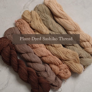 May include: A collection of six braided skeins of plant-dyed Sashiko thread arranged in a diagonal row, featuring a gradient of earth tones ranging from dark brown to light beige, with the text Plant-Dyed Sashiko Thread overlaid in the centre.