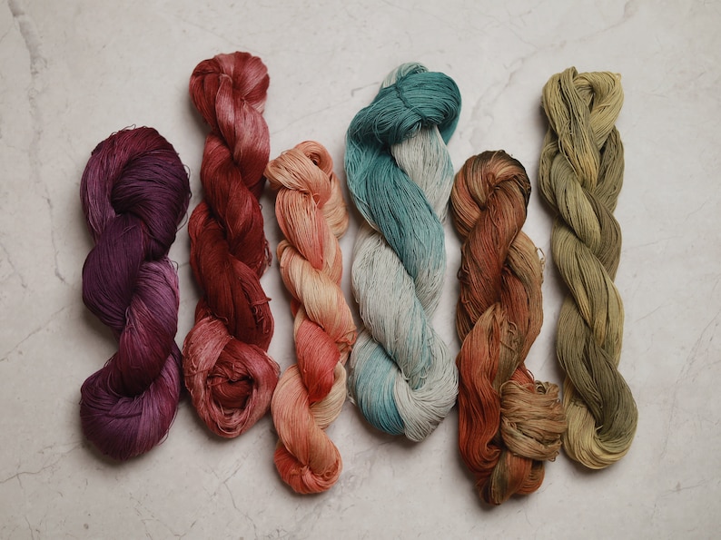 Puede incluir: Seis madejas de hilo trenzado dispuestas en fila, con una variedad de colores que incluyen morado intenso, rojo oscuro, melocot&oacute;n claro, verde azulado y blanco, &oacute;xido y marr&oacute;n matizado, y verde oliva apagado, cada una con textura.
