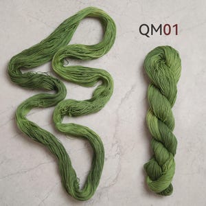 Puede incluir: Una madeja de hilo verde oliva presentada en dos formas: una parte enrollada de forma suelta en una figura alargada y sinuosa, y otra parte retorcida en un paquete compacto. El texto QM01 est&aacute; impreso en rojo oscuro arriba a la derecha.