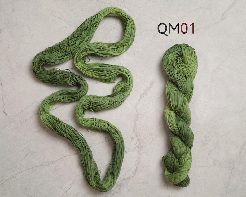 Puede incluir: Una madeja de hilo verde oliva presentada en dos formas: una parte enrollada de forma suelta en una figura alargada y sinuosa, y otra parte retorcida en un paquete compacto. El texto QM01 est&aacute; impreso en rojo oscuro arriba a la derecha.