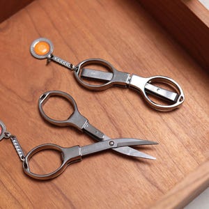Könnte beinhalten: Zwei Paar silberfarbene, vielseitige Scheren mit orangefarbenen und roten Akzenten. Die Scheren haben einen Ring am Ende des Griffs, so dass sie an einem Schlüsselbund befestigt werden können.
