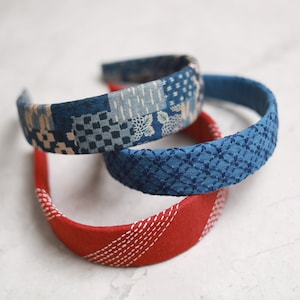 Peut inclure: Trois bandeaux pour cheveux en tissu, dans les tons de bleu et de rouge. Le bandeau supérieur présente un motif patchwork avec des motifs floraux et géométriques. Le bandeau du milieu est bleu uni avec un motif cousu. Le bandeau inférieur est rouge avec des coutures blanches.