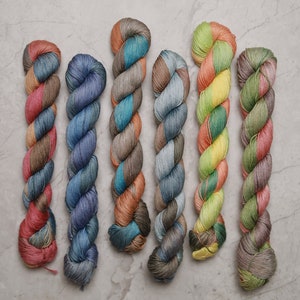 May include: Six skeins of hand-dyed yarn in various colour combinations, including shades of blue, green, yellow, orange, and brown. The yarn is twisted and arranged in a vertical line.