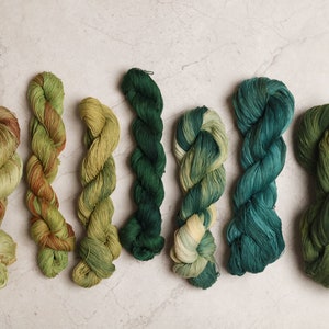 Puede incluir: Siete madejas de hilo teñido a mano en varios tonos de verde, que van desde el oliva claro hasta el verde bosque profundo. El hilo está dispuesto en una fila, mostrando las variaciones de color y las texturas. Las madejas están bien enrolladas y parecen suaves.