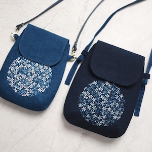 Pode incluir: Duas bolsas crossbody azul índigo com fechos de aba. Cada bolsa apresenta um design circular bordado com padrões florais brancos e azuis. As bolsas têm alças ajustáveis e são exibidas em uma superfície cinza claro.