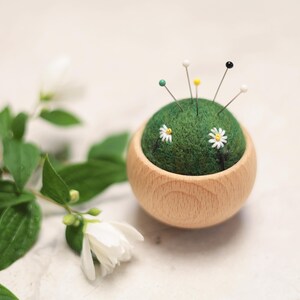 May include: A round wooden pincushion with a green felt top, decorated with embroidered daisies and topped with colorful pins. A sprig of white flowers and green leaves is in the background, creating a natural, handcrafted aesthetic.