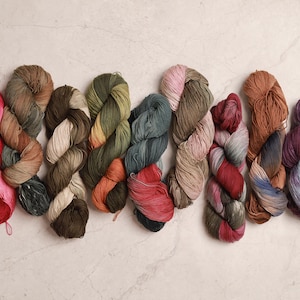 May include: Ten skeins of yarn in various colours, including red, pink, green, brown, and purple. The yarn is arranged in a row on a white marble surface.