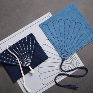 Peut inclure: Une enveloppe en tissu bleu foncé avec des coutures sashiko blanches et un cordon blanc, un éventail en tissu bleu clair avec des coutures sashiko bleu foncé et un cordon bleu foncé, et un pochoir blanc avec un motif d'éventail noir.