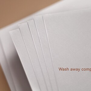 May include: A stack of white paper sheets with a soft texture, fanned out to reveal the edges. The text "Wash away completely" is printed in brown at the bottom right corner, set against a blurred brown background.