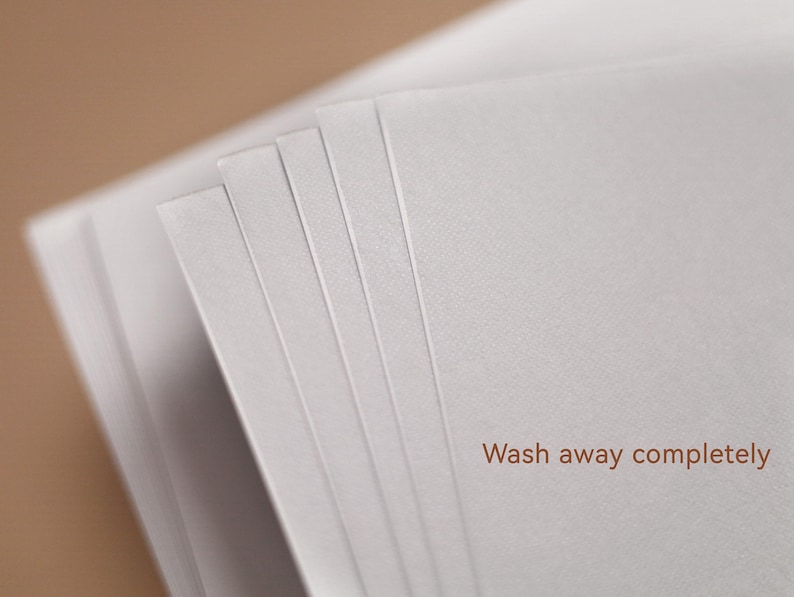 May include: A stack of white paper sheets with a soft texture, fanned out to reveal the edges. The text "Wash away completely" is printed in brown at the bottom right corner, set against a blurred brown background.