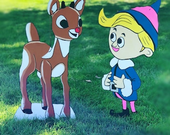 May include: A flat, cutout-style yard decoration featuring a brown reindeer with a bright red nose standing next to a cartoon elf. The elf has yellow hair, a blue hat with a pink tip, a blue jacket with white trim, and pink leggings.