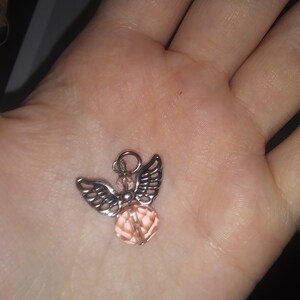 May include: A small, silver-toned angel pendant with detailed wings and a faceted, light pink crystal body. The pendant has a small loop at the top for hanging. The angel is displayed against a blurred background.