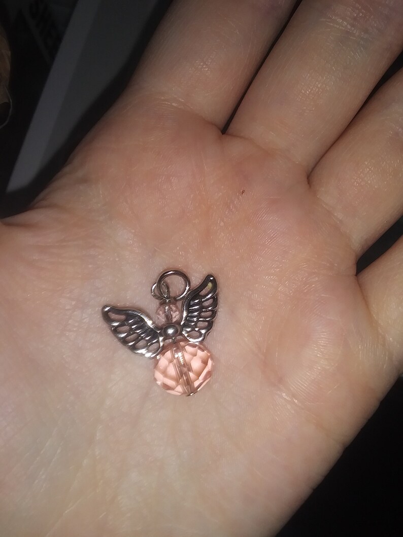 May include: A small, silver-toned angel pendant with detailed wings and a faceted, light pink crystal body. The pendant has a small loop at the top for hanging. The angel is displayed against a blurred background.
