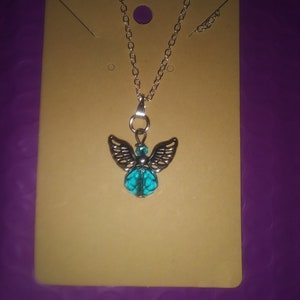 May include: Silver-toned necklace featuring an angel pendant with detailed wings and a turquoise-colored bead. The necklace is displayed on a tan card against a purple background. The chain is delicate and the pendant is the focal point.