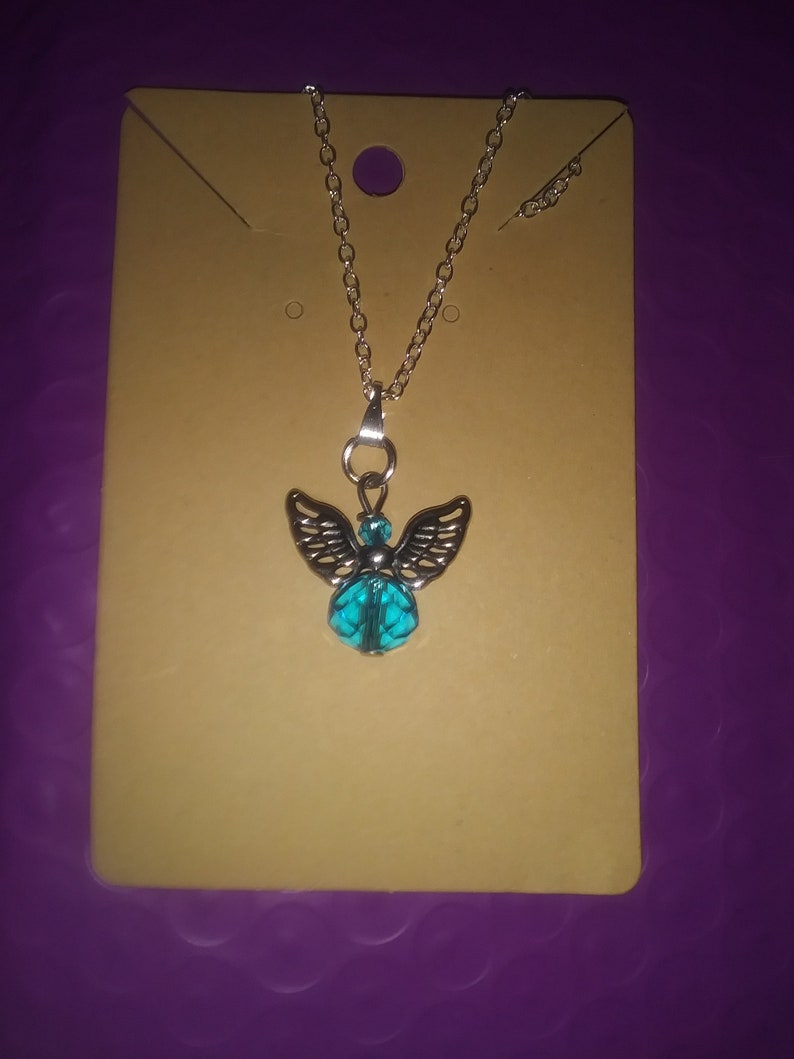 May include: Silver-toned necklace featuring an angel pendant with detailed wings and a turquoise-colored bead. The necklace is displayed on a tan card against a purple background. The chain is delicate and the pendant is the focal point.