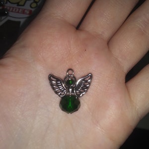 May include: A silver-toned angel pendant with detailed wings and a faceted, emerald-green crystal body. The pendant is designed with a small loop for hanging and is held in an open palm.