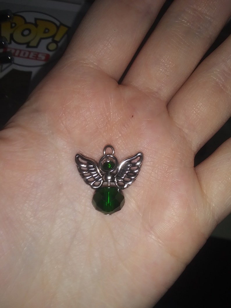 May include: A silver-toned angel pendant with detailed wings and a faceted, emerald-green crystal body. The pendant is designed with a small loop for hanging and is held in an open palm.
