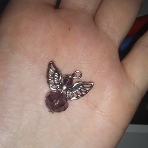 May include: A small, silver-toned angel pendant with a faceted, purple bead body and detailed wing design. The pendant is held in a palm, showcasing its delicate features and craftsmanship. The angel charm is designed for jewelry making.