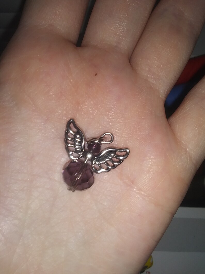 May include: A small, silver-toned angel pendant with a faceted, purple bead body and detailed wing design. The pendant is held in a palm, showcasing its delicate features and craftsmanship. The angel charm is designed for jewelry making.