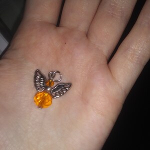 May include: A small, silver-toned angel charm with orange crystal bead accents. The angel has detailed wings and a round, faceted body. The charm is held in an open hand.