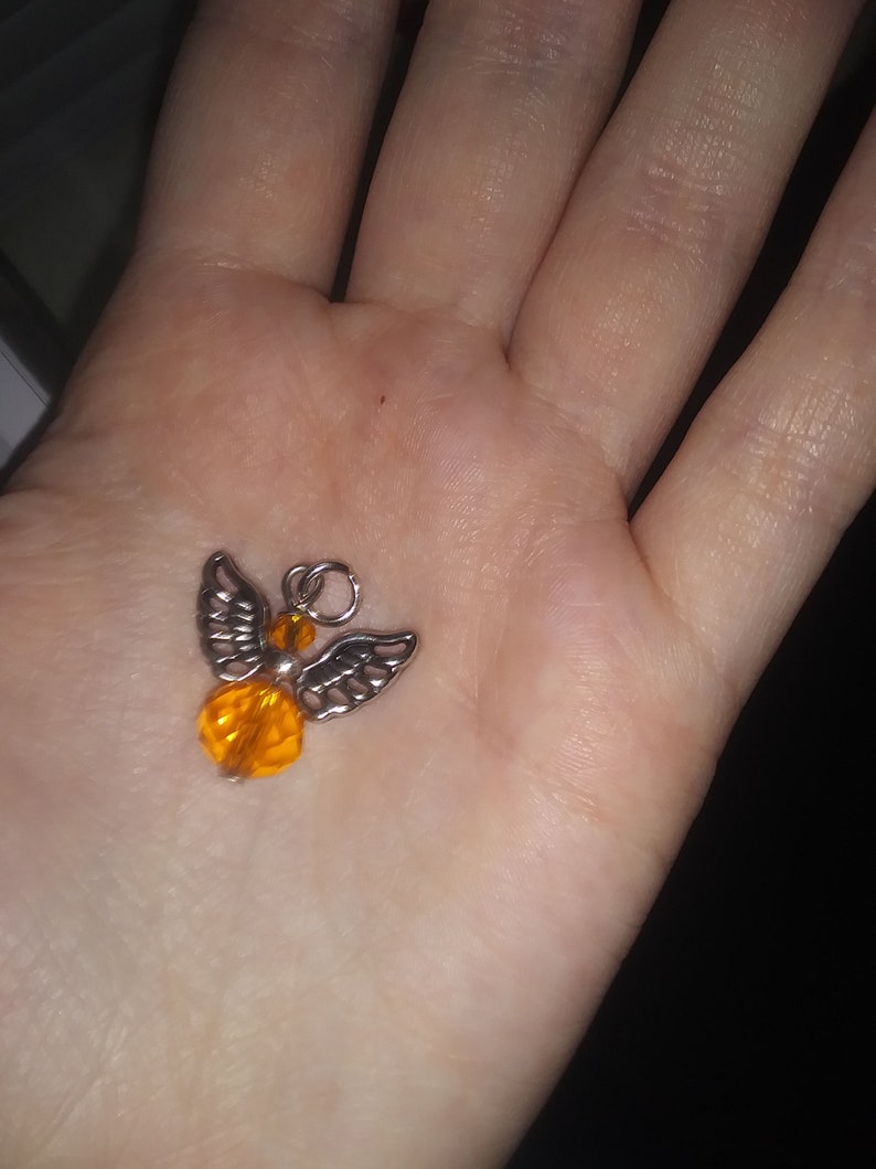 May include: A small, silver-toned angel charm with orange crystal bead accents. The angel has detailed wings and a round, faceted body. The charm is held in an open hand.