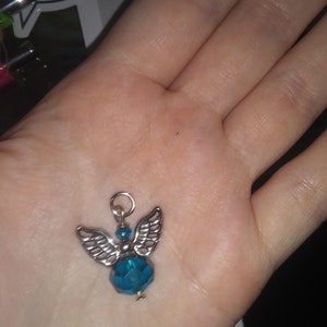 May include: A small angel charm with silver-colored wings and a teal-colored faceted bead body. The charm has a silver loop at the top for attaching to a necklace or bracelet. The charm is held in a person's hand.