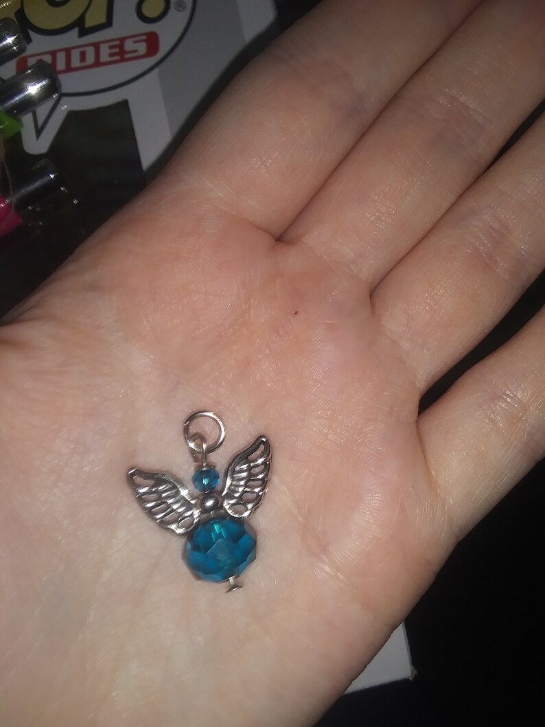 May include: A small angel charm with silver-colored wings and a teal-colored faceted bead body. The charm has a silver loop at the top for attaching to a necklace or bracelet. The charm is held in a person's hand.