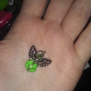May include: A small angel charm with silver-colored wings and a faceted green bead body. The charm is held in a hand, showcasing its delicate details and design. The angel charm is a decorative accessory.
