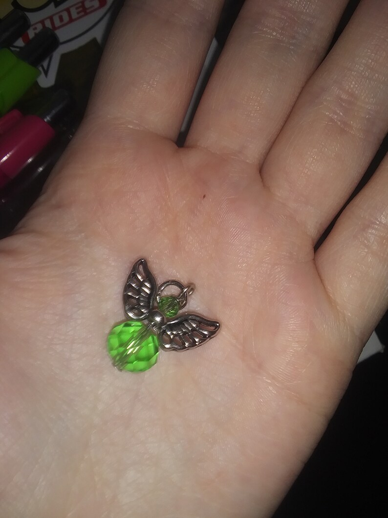 May include: A small angel charm with silver-colored wings and a faceted green bead body. The charm is held in a hand, showcasing its delicate details and design. The angel charm is a decorative accessory.
