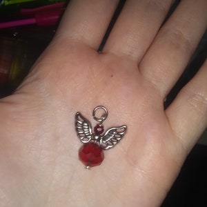 May include: A small, silver-toned angel charm with detailed wings and a faceted, deep red crystal body. The charm has a small loop at the top for attaching to a necklace or bracelet. The angel is a decorative jewelry element.