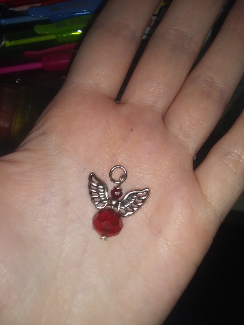 May include: A small, silver-toned angel charm with detailed wings and a faceted, deep red crystal body. The charm has a small loop at the top for attaching to a necklace or bracelet. The angel is a decorative jewelry element.