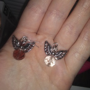 May include: A pair of angel-shaped earrings. Each earring features silver-toned wings and a faceted, teardrop-shaped bead. One bead is a reddish-brown color, and the other is a light peach color. The earrings are displayed on a hand.