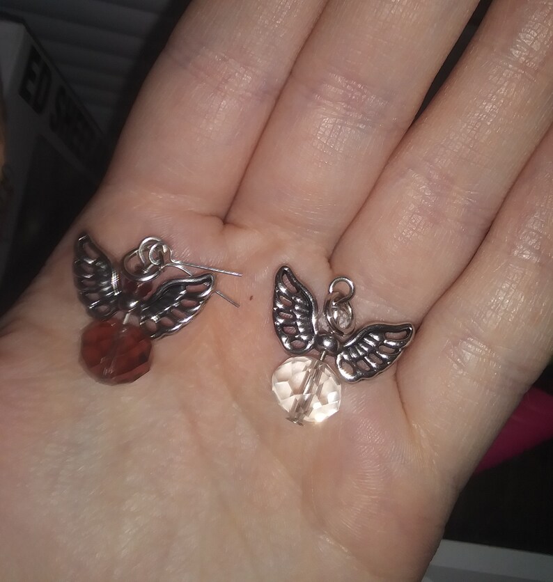May include: A pair of angel-shaped earrings. Each earring features silver-toned wings and a faceted, teardrop-shaped bead. One bead is a reddish-brown color, and the other is a light peach color. The earrings are displayed on a hand.