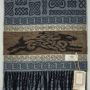 Può includere: Una sciarpa tessuta blu e marrone con un motivo a nodo celtico e una frangia nera. La sciarpa ha una striscia marrone con un motivo a nodo celtico nero al centro. La sciarpa ha un'etichetta che dice "Calzeat Celtic Collection".