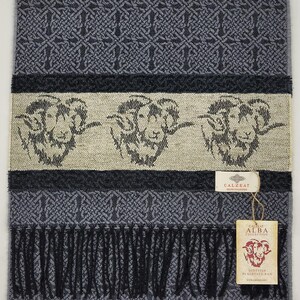May include: A black and grey patterned scarf with a fringe. The scarf has a beige stripe with three black ram heads. The scarf has a tag that says "Calzeat Alba Collection Scottish Blackface Ram".